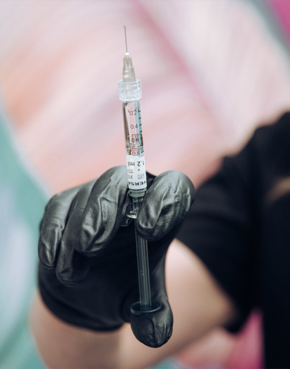 A gloved hand holds a syringe filled with a clear liquid. The syringe is positioned vertically, with the needle pointing upwards. The background is blurry and appears to be a colorful wall.